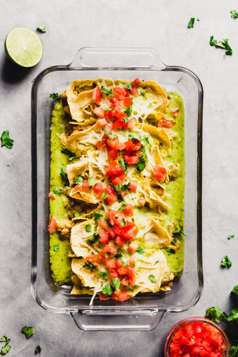 a casserole dish of white bean enchiladas served alongside a half a lime and a bowl of diced tomatoes