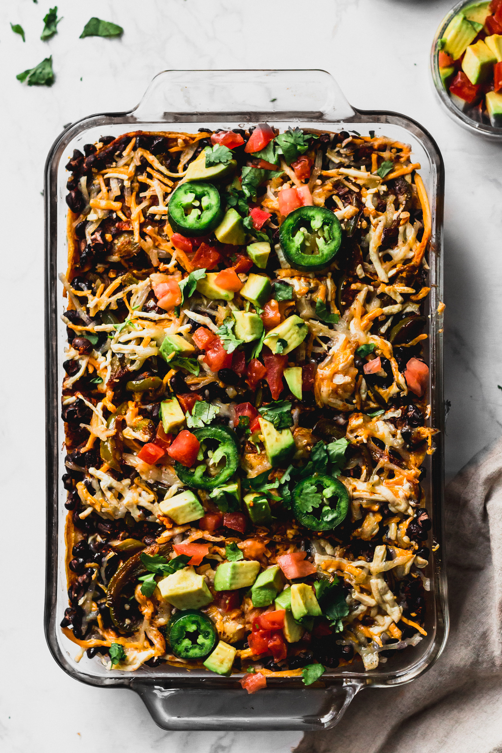 a casserole dish filled with vegan enchiladas topped with tomatoes, avocado, jalapenos and vegan cheese