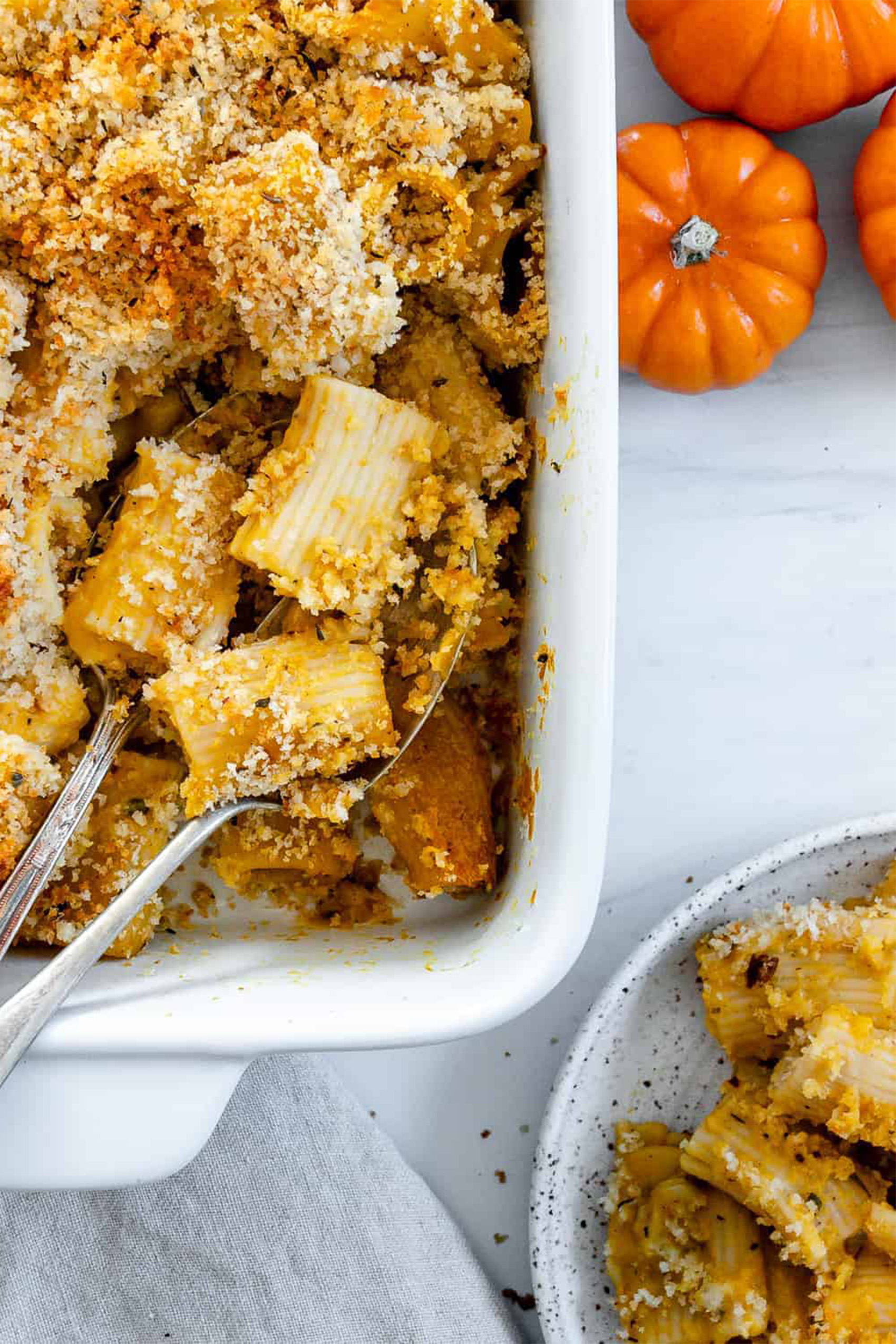 a spoon scooping into a baking dish filled with vegan pumpkin pasta that has been topped with breadcrumbs