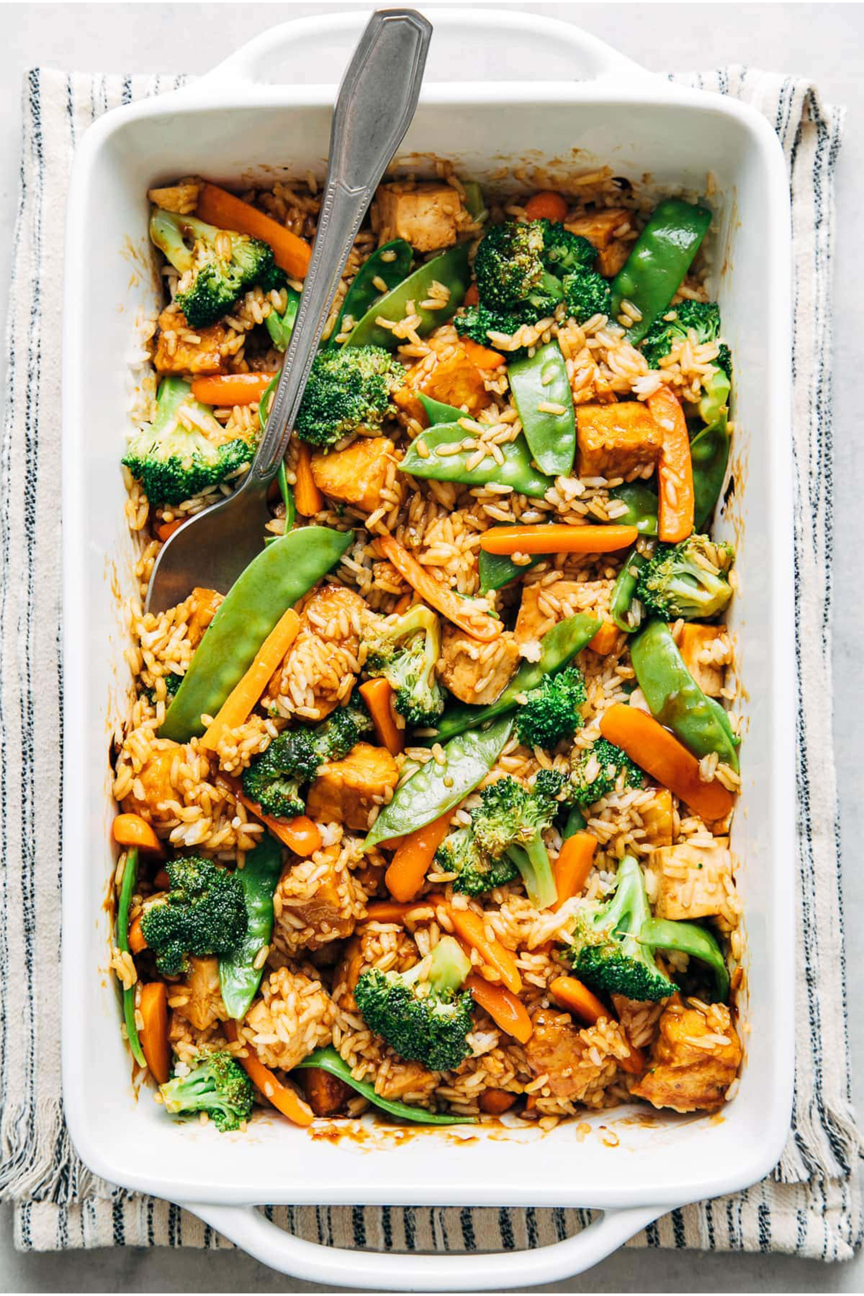 a casserole dish filled with rice, tempeh, broccoli, green peas, peppers and carrots