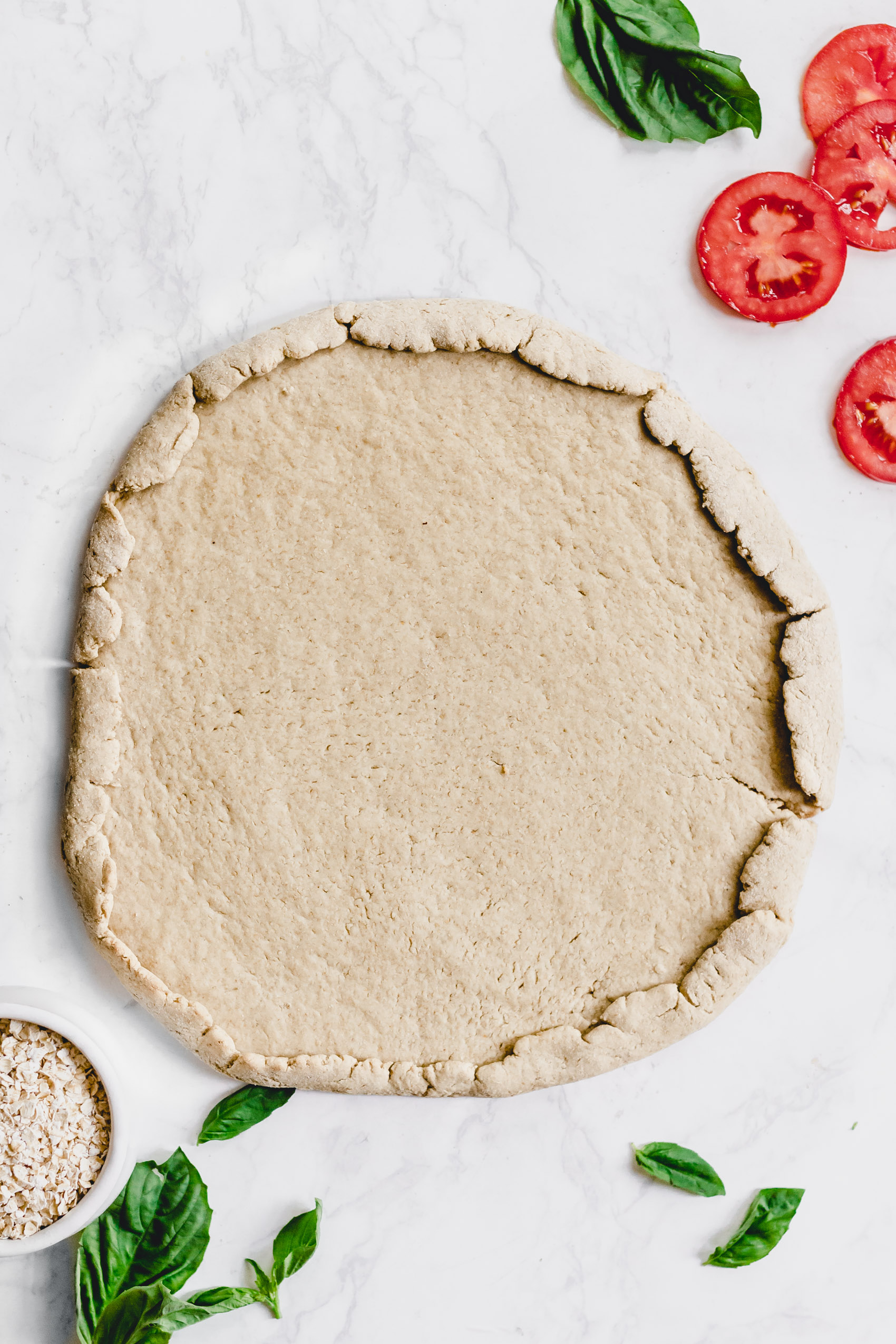 a round homemade pizza crust next to sliced tomatoes