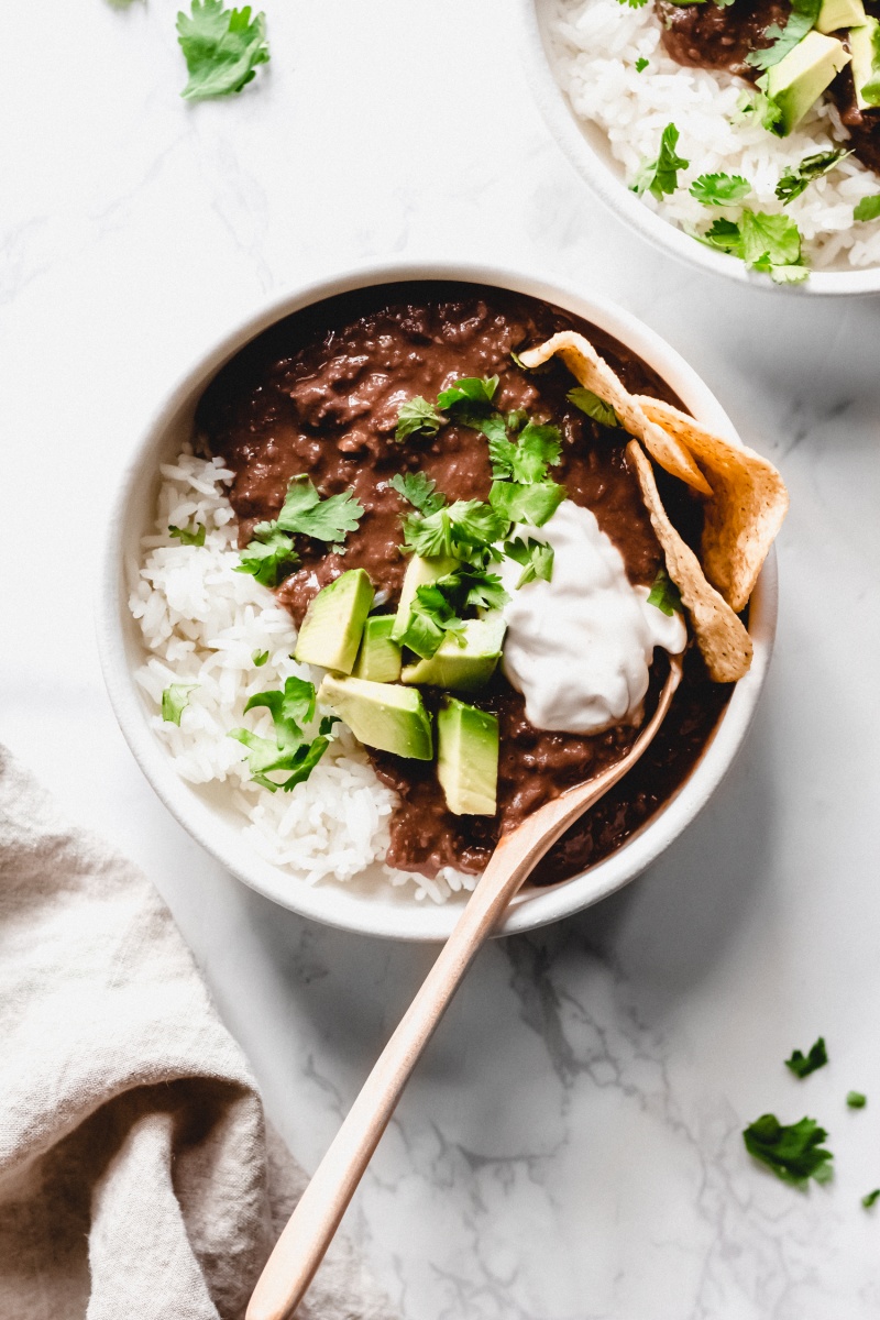 a bowl of black bean soup topped with cilantro, sour cream, and avocado served with chips and rice
