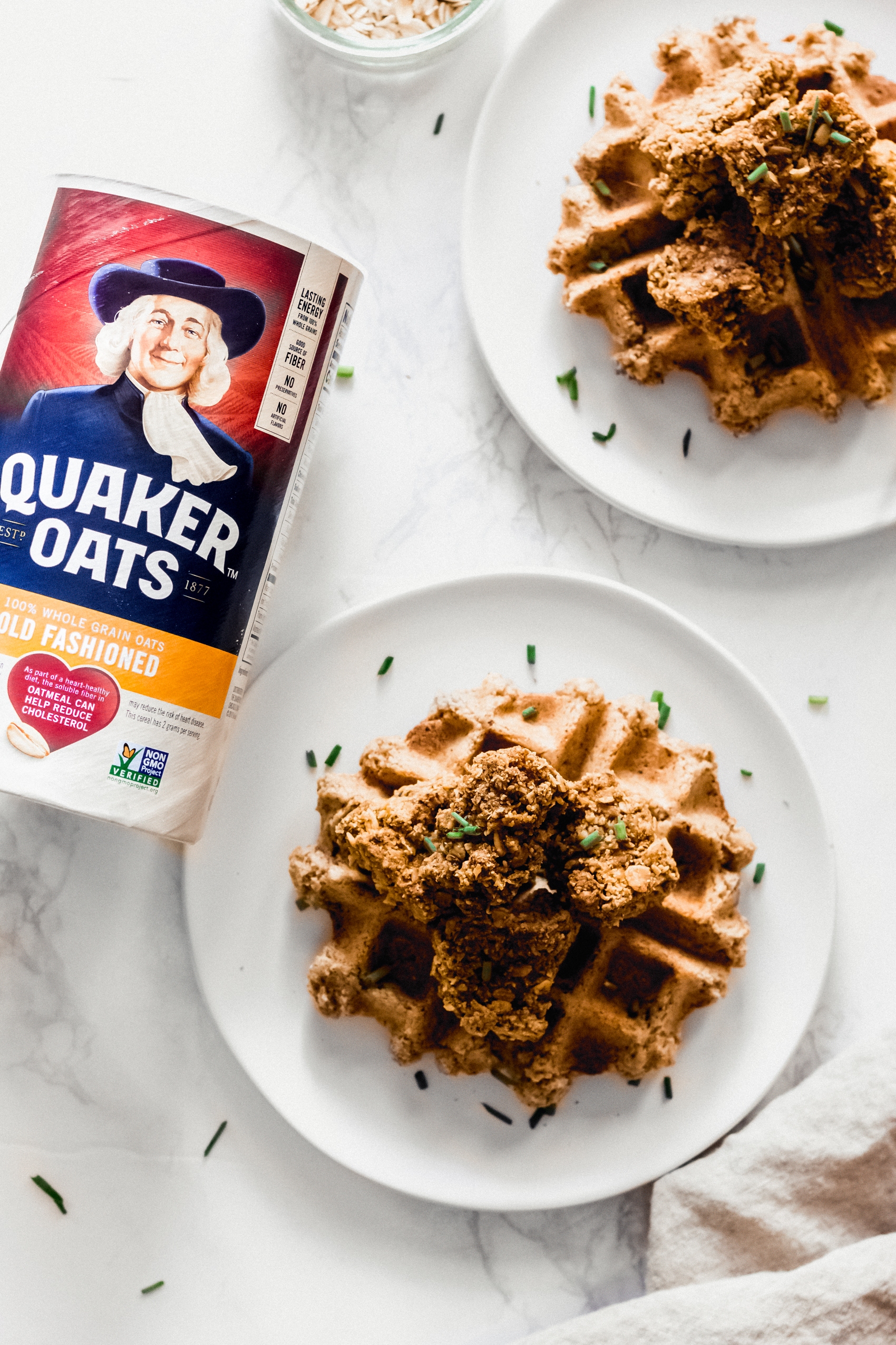 two plates of vegan chicken and waffles sit next to a container of oats