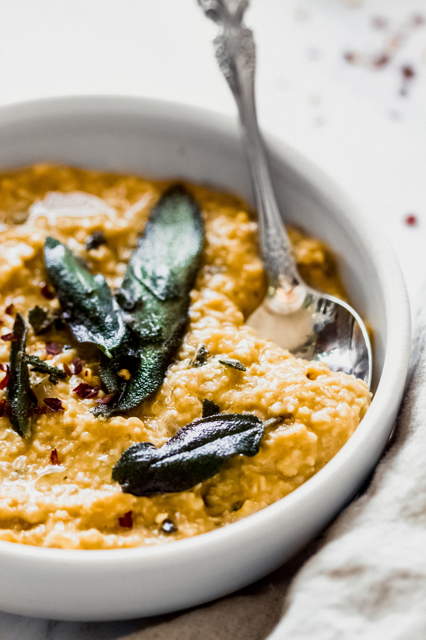 a spoon taking a scoop of pumpkin savory oatmeal topped with chili flake and sage leaves