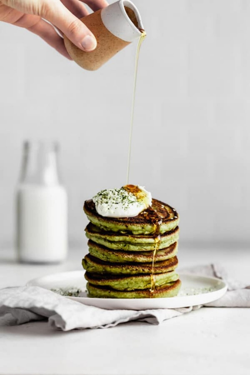 stack of green pancakes on plate with white sauce on top and maple syrup poured on 