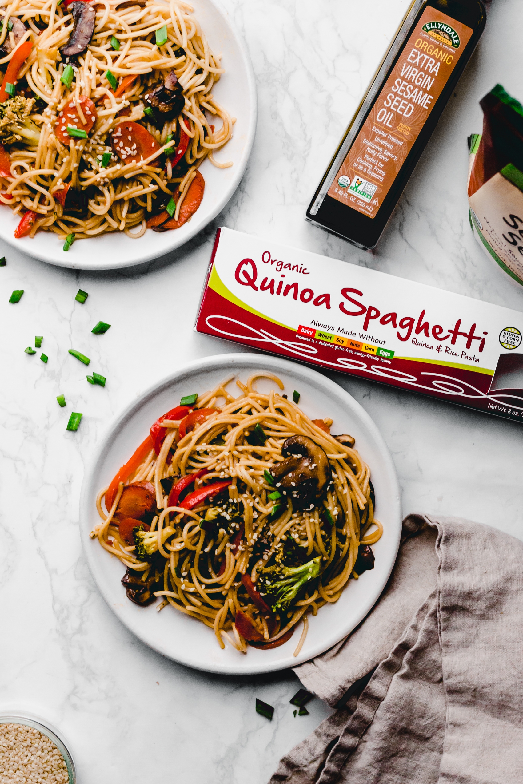 two plates of vegetable lo mein served alongside a box of gluten-free spaghetti and a bottle of sesame seed oil