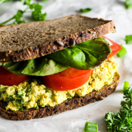 a vegan egg salad sandwich resting on parchment paper surrounded by fresh parsley