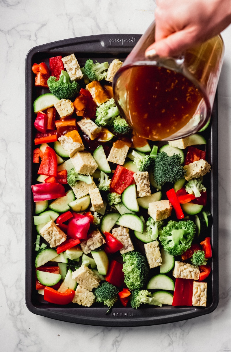 a measuring cup of vegan teriyaki sauce is poured over a sheet tray of veggies and tempeh