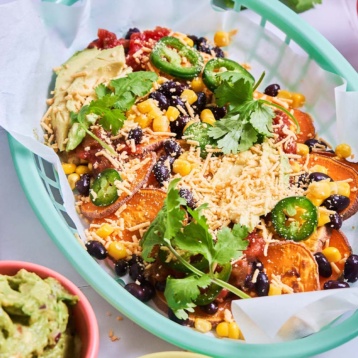 a basket of vegan sweet potato nachos topped with cheese, jalapenos, cilantro, black beans and corn served with a side of guacamole