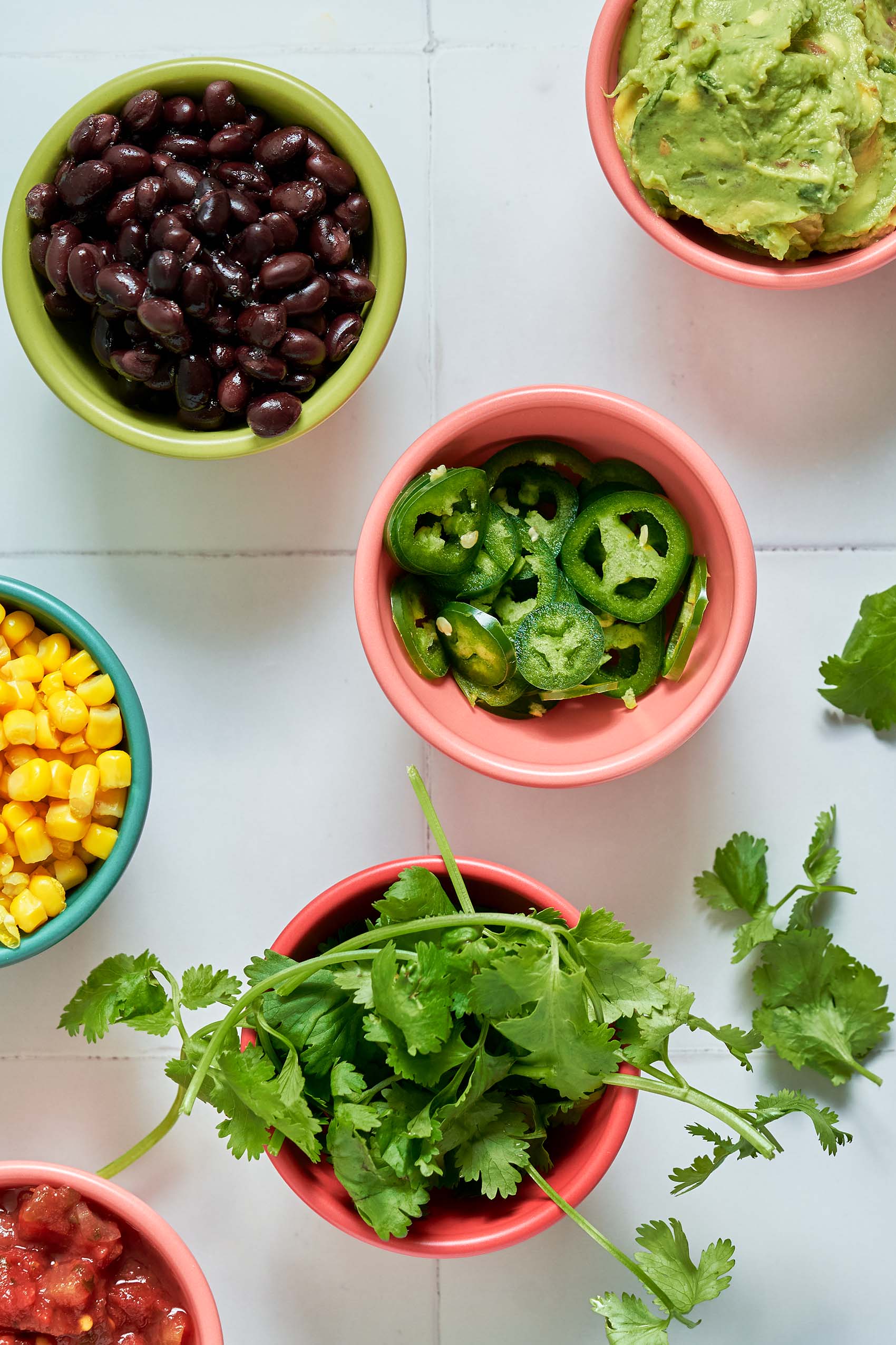 bowls of nacho toppings including sliced jalapenos, black beans, cilantro, corn, salsa and guacamole