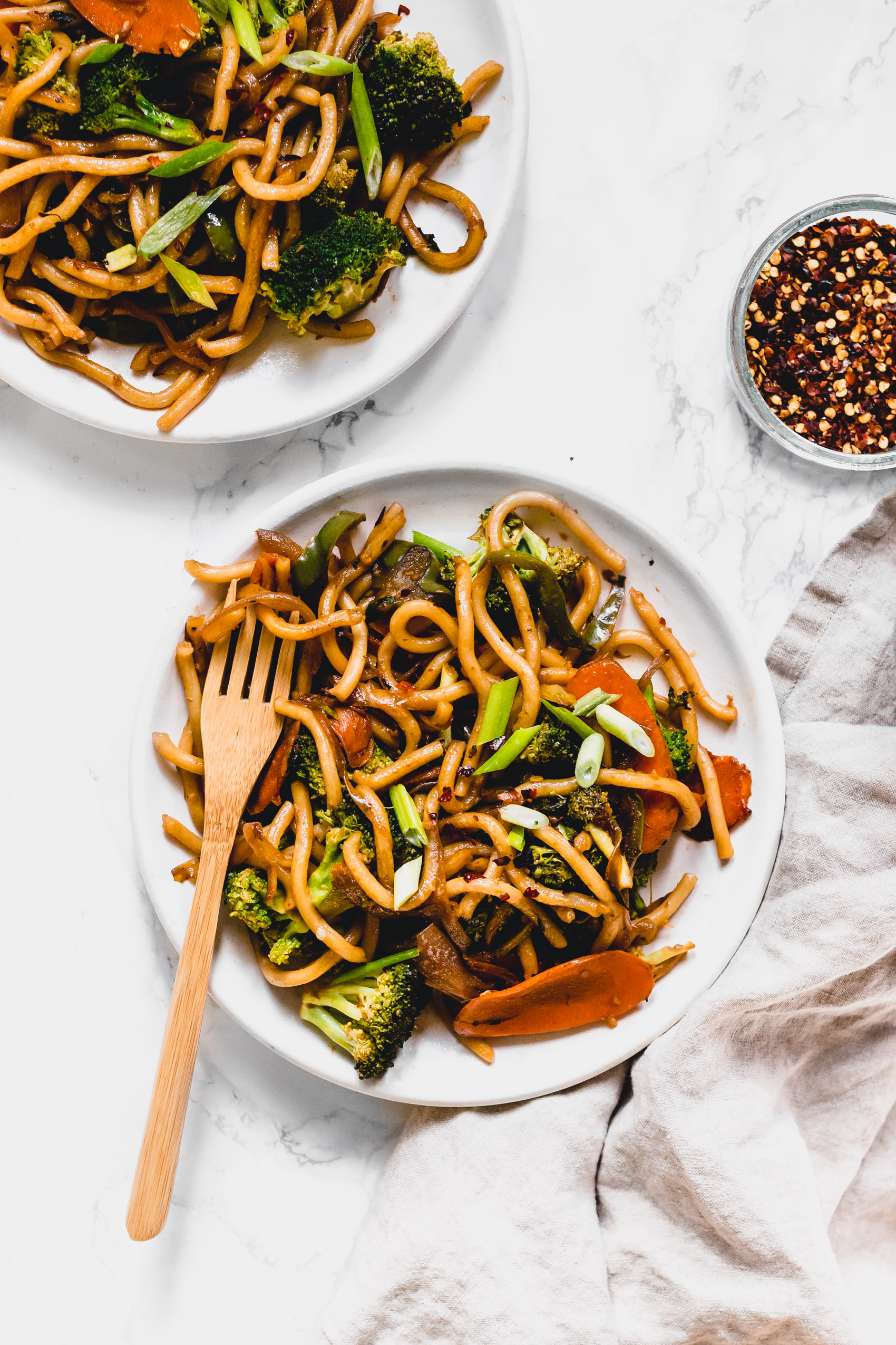 two plates of vegan drunken noodles next to a small bowl of dried chili flakes