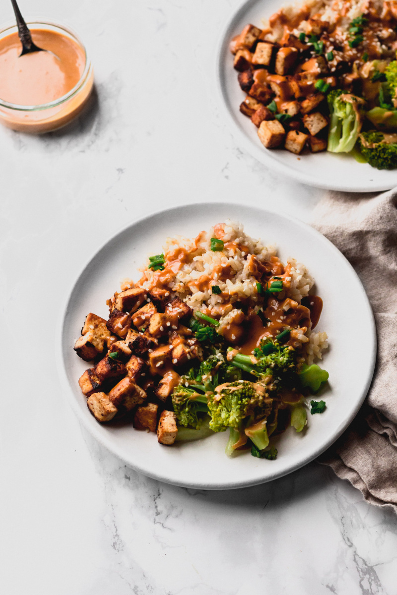 side view of two plates with brown grains, crispy fried tofu, and steamed broccoli drizzled with peanut sauce