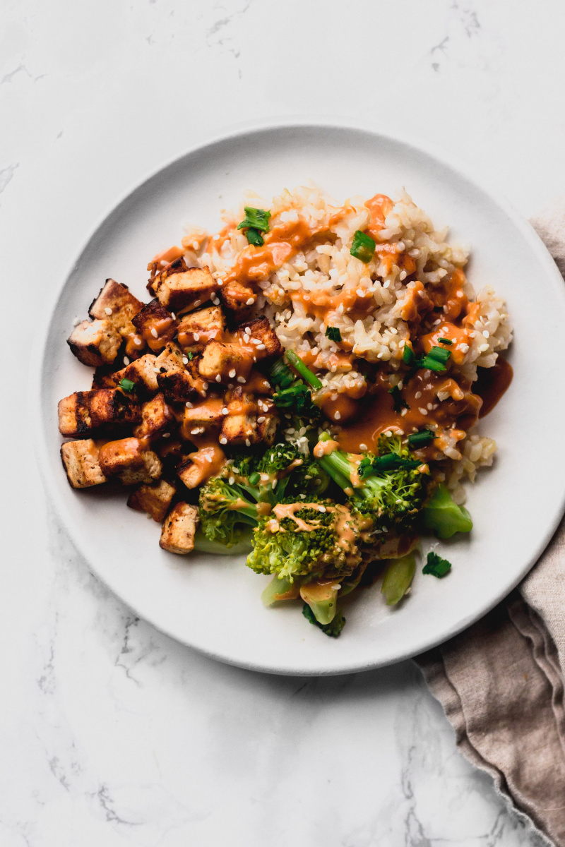 close up of a brown rice bowl with fried tofu, broccoli, peanut sauce, sesame seeds, and green onions