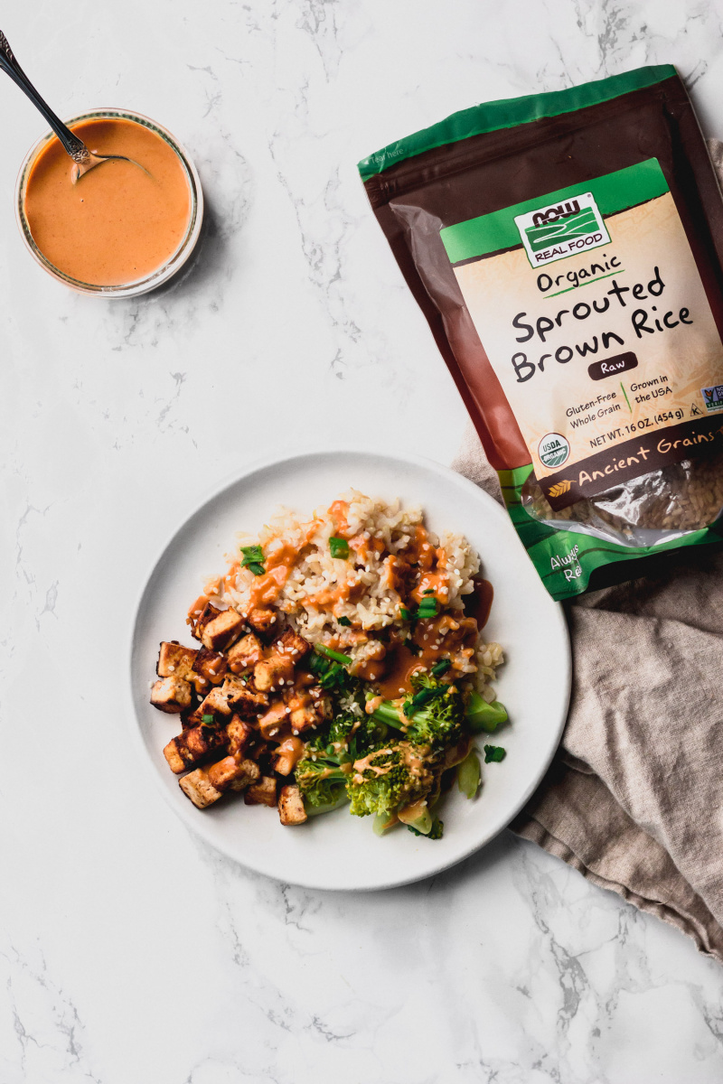 overhead view of a plate with tofu, rice, and broccoli next to a bag of brown rice and a bowl of peanut sauce