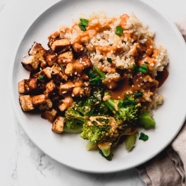 close up of a brown rice bowl with fried tofu, broccoli, peanut sauce, sesame seeds, and green onions