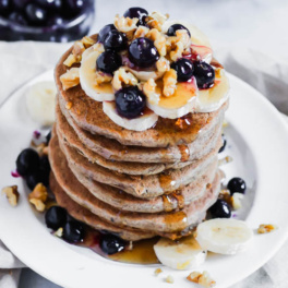 a stack of vegan buckwheat pancakes topped with nuts, bananas, blueberries and maple syrup