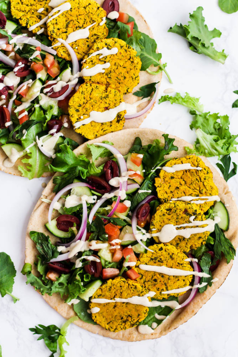 Two pitas topped with lettuce, olives, cucumber slides, red onion, chopped tomatoes and four turmeric falafels each. The pita sandwiches are topped with a tahini drizzle