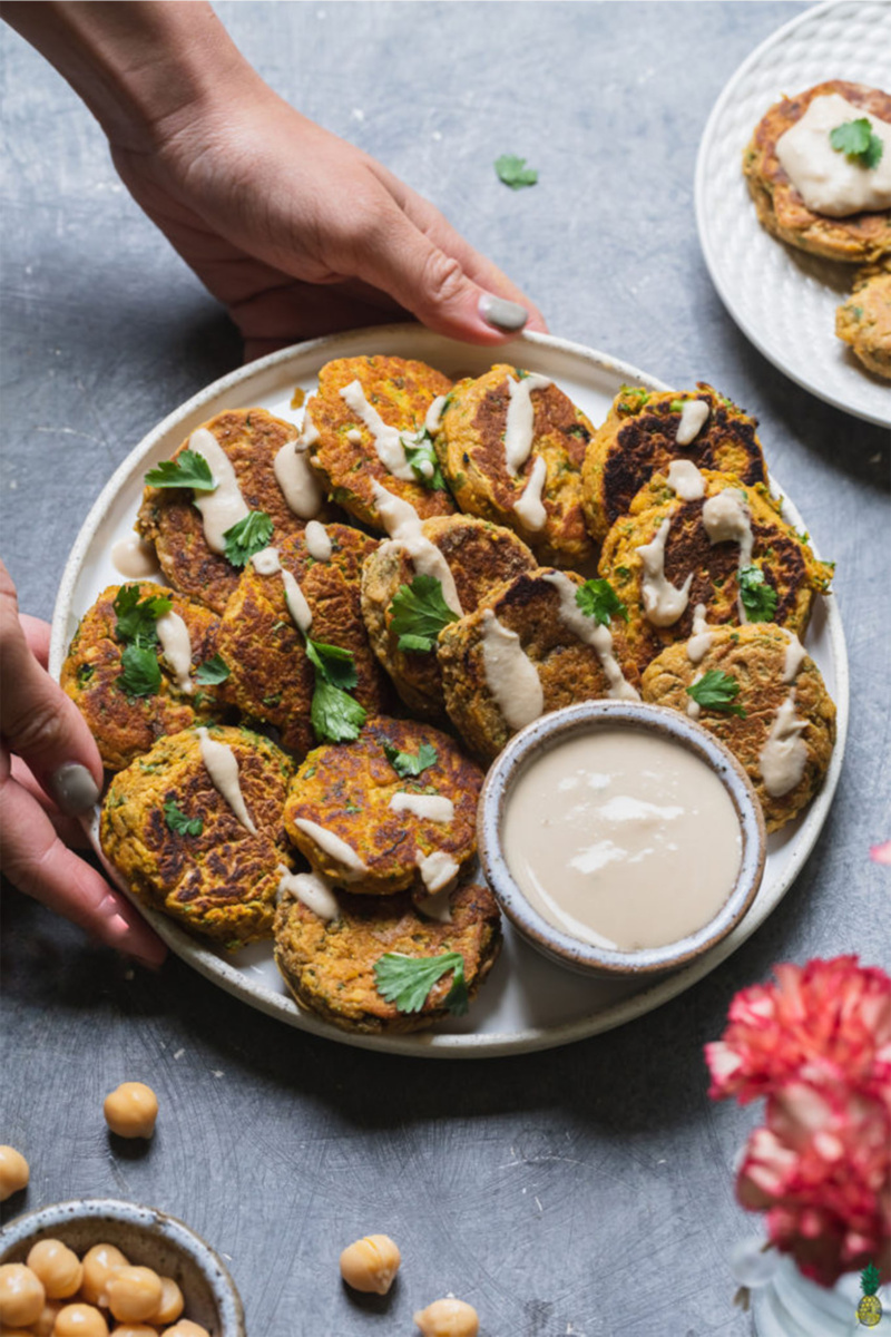 Two hands serving a plate of pumpkin chickpea fritters. The fritters are topped with cilantro and served with hummus