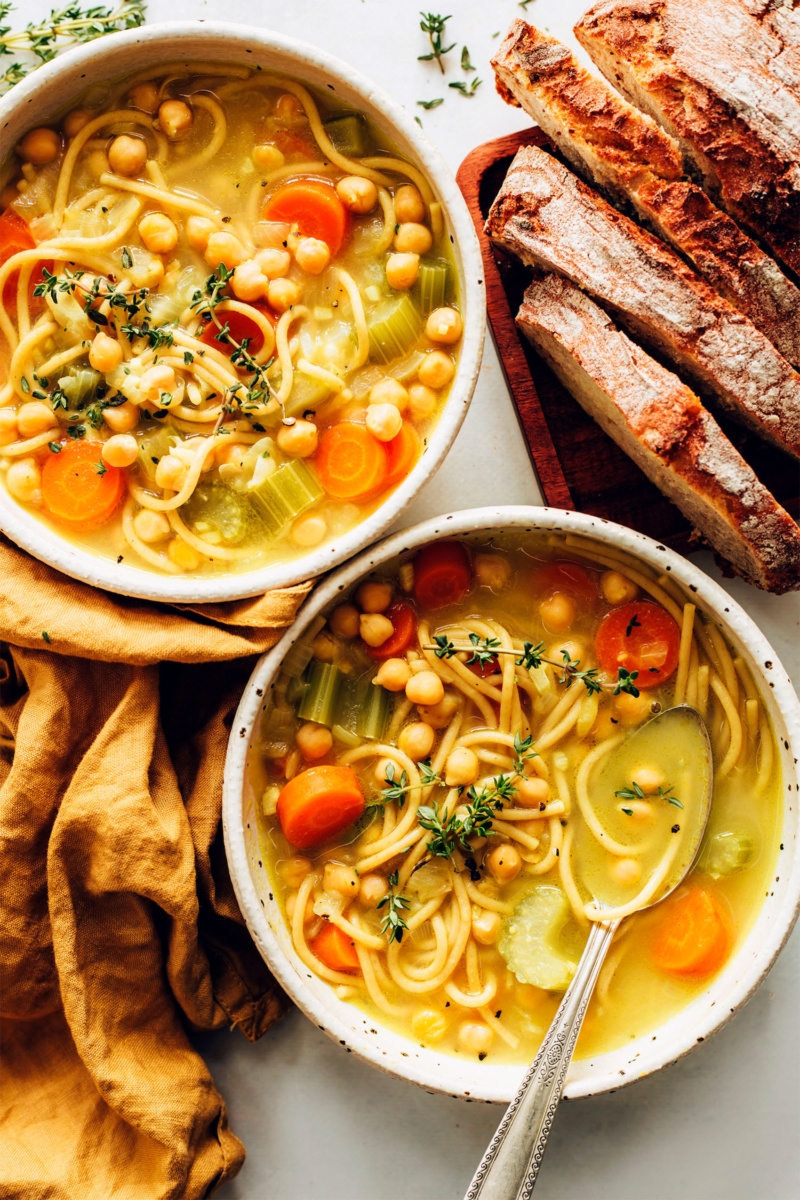 Two bowls of chickpea noodle soup served alongside slices of a crusty white bread