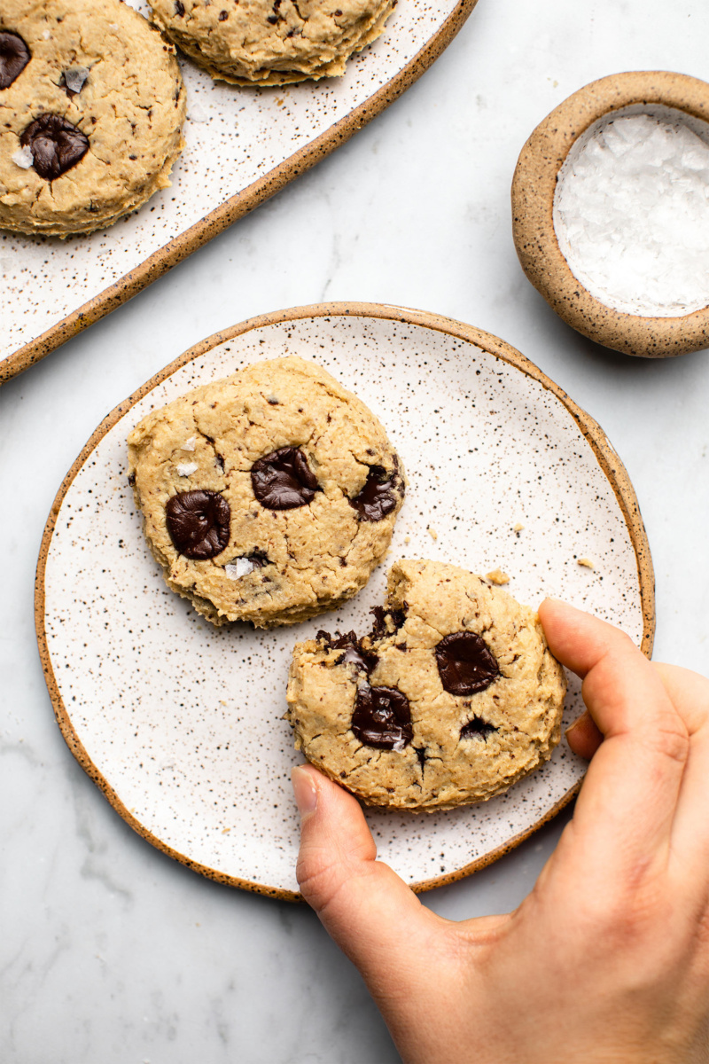 Two chickpea chocolate chip cookies served on a small white plate. One cookie has a bite taken out of it and a larger dish of cookies can be seen in the top left corner of the image