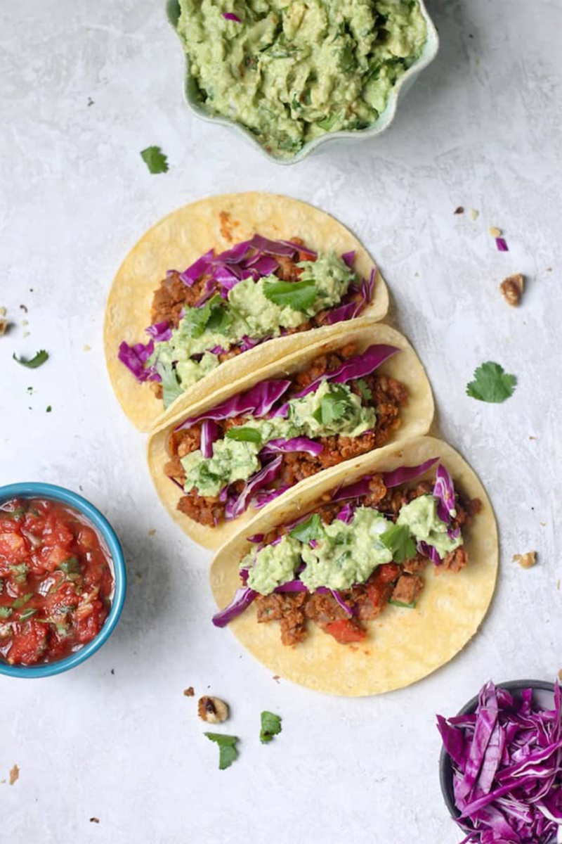 Three vegan walnut and chickpea tacos on a gray stone countertop. The tacos are topped with guacamole and purple cabbage. A bowl of guacamole, a bowl of red cabbage and a bowl of salsa surround the tacos