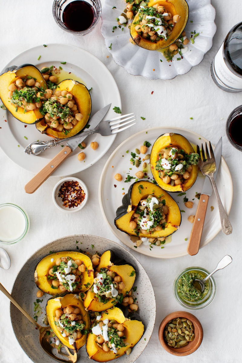 An overhead shot of four plates, each with servings of quartered acorn squashes stuffed with chickpeas, chimichurri and some topped with yogurt. Bowls of chili flakes, pumpkin seeds, chimichurri and yogurt along with two glasses of red wine surround the plates