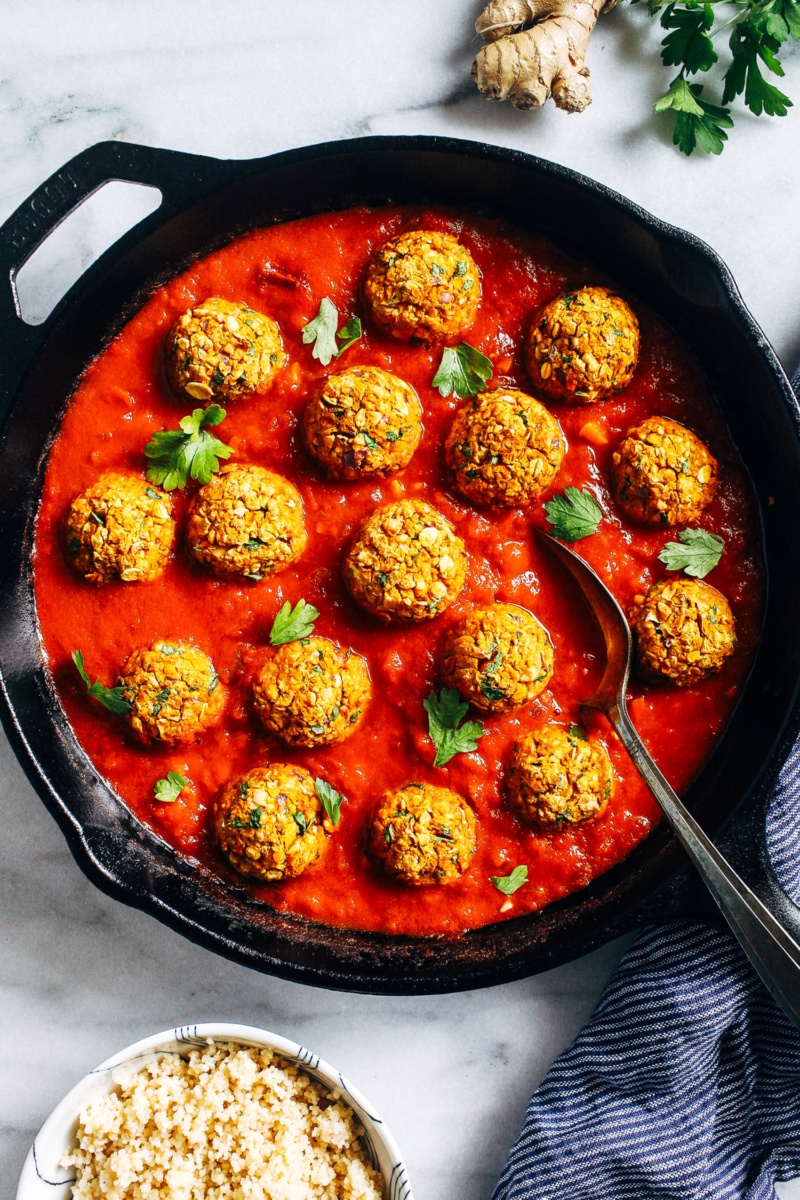 A cast iron skillet serving a red Moroccan sauce and chickpea meatballs. The sauce is topped with sprigs of cilantro. A small bowl of quinoa, a knob of ginger and a spring of cilantro surround the skillet