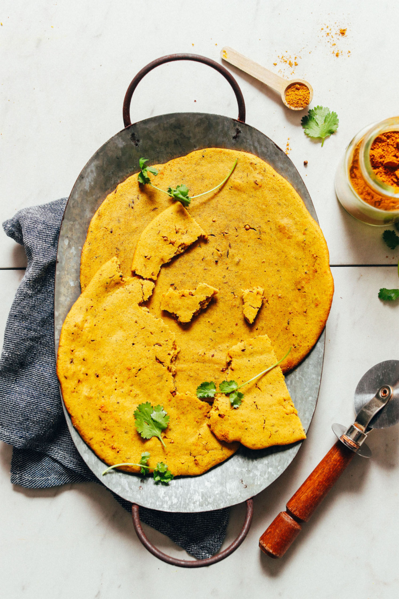 A tin platter holding two pieces of socca, or chickpea bread. One pieces of socca has been torn into pieces. A small pizza cutter and jar of turmeric can be seen on the right side of the image