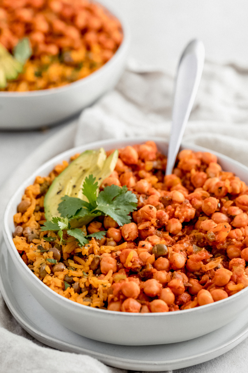 A bowl of chickpeas in sofrito served with arroz con grandules. The dish is topped with avocado slices and a sprig of cilantro