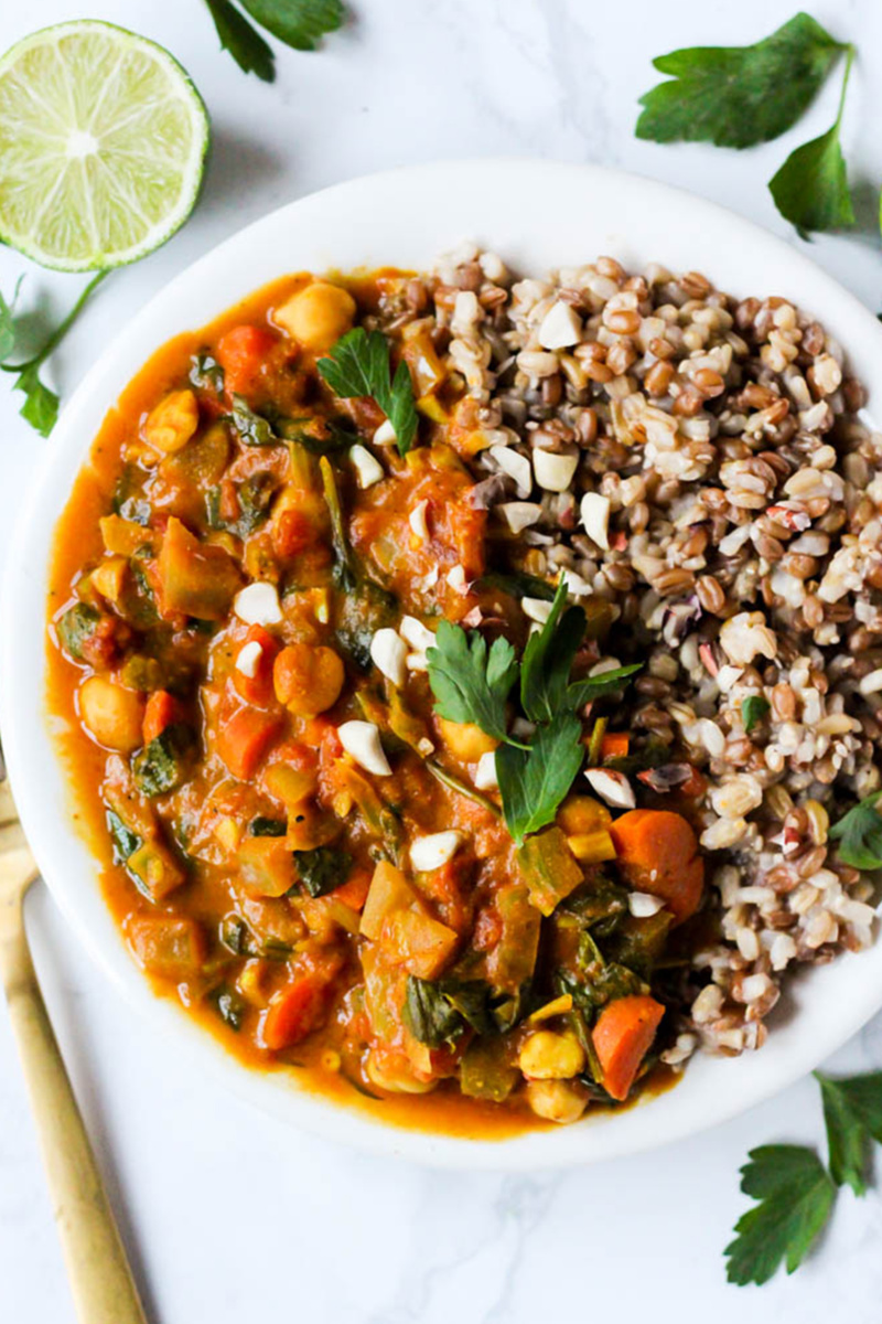 A white bowl filled with chickpea peanut stew on the left and a serving of grains on the right. The bowl is surrounded by sprigs of parsley and a half a lime