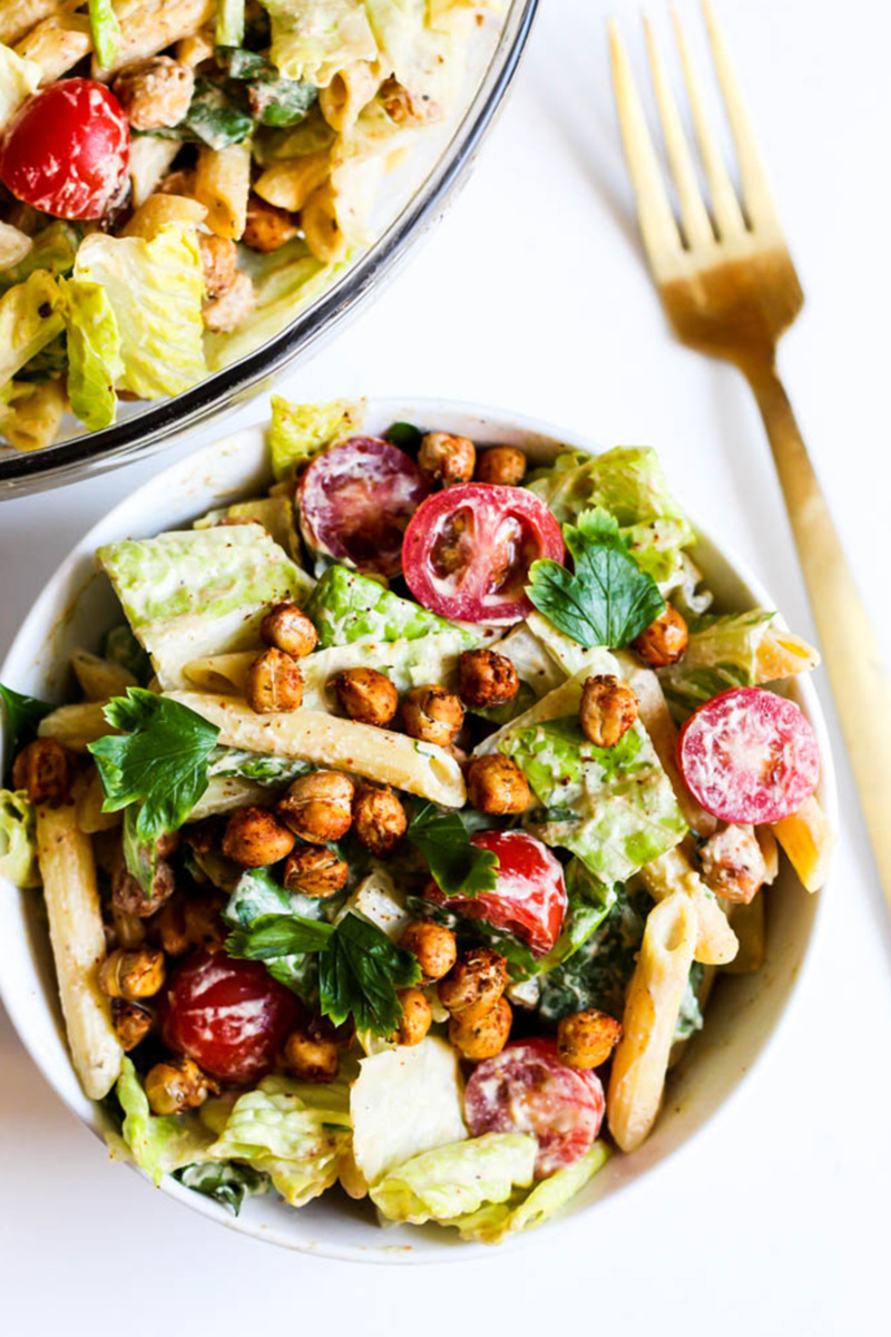 A bowl of pasta salad consisting of romaine, penne pasta, cherry tomatoes, caesar dressing and crispy chickpeas