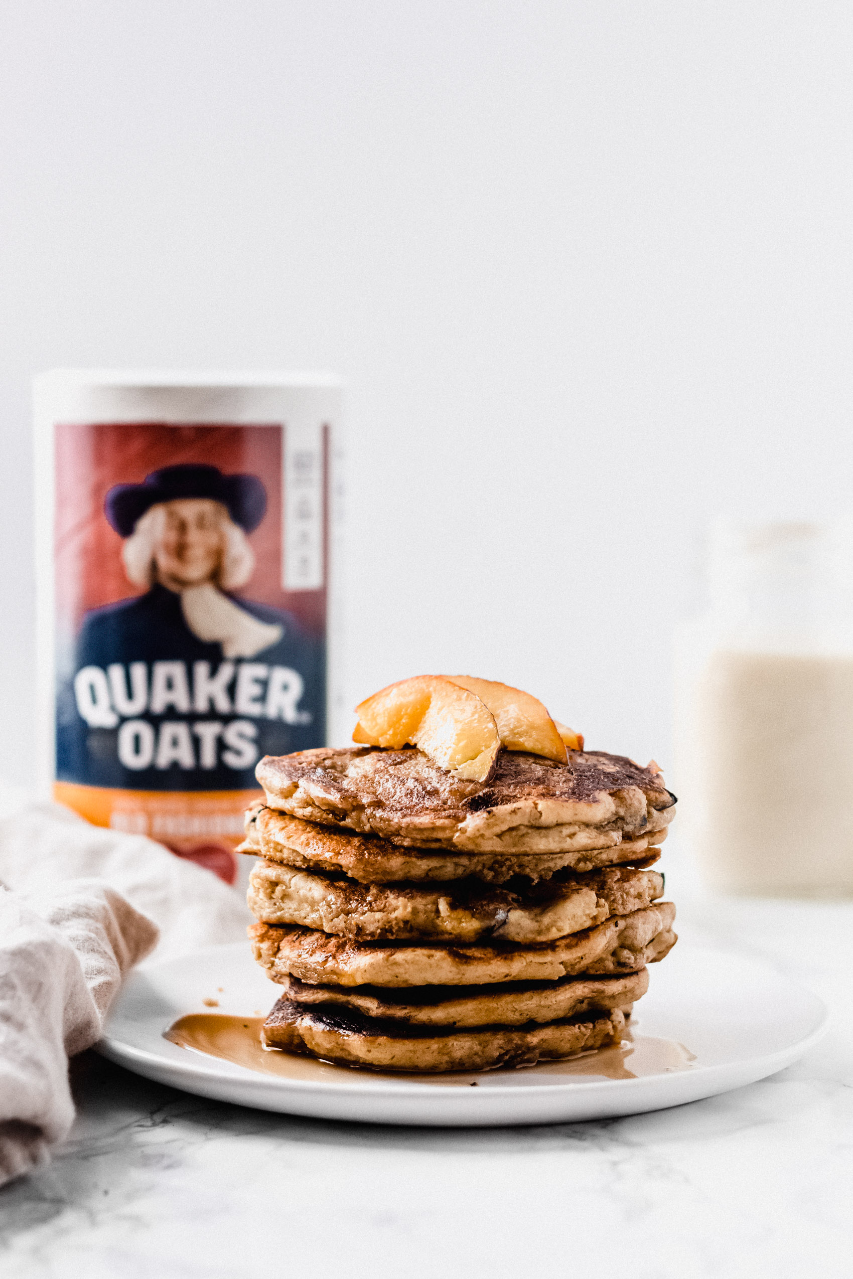 a stack of pancakes with sliced peaches and maple syrup on top with a carton of oats behind them