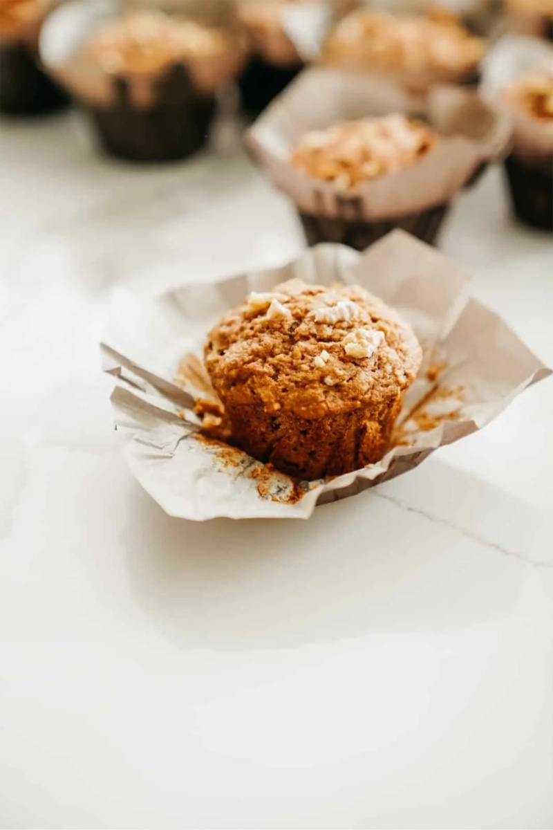 a vegan apple cinnamon muffin with the paper peeled back sits on a white countertop