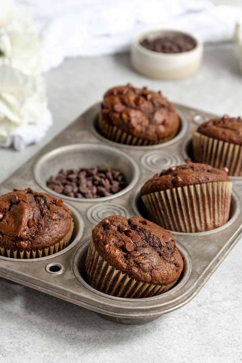 A muffin tray holds 5 double chocolate muffins, with one muffin slot filled with chocolate chips