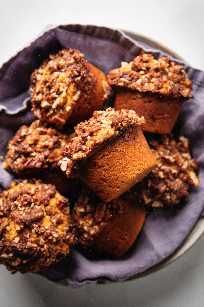 A bowl lined with a purple towel is piled high with sweet potato muffins topped with a pecan crumble