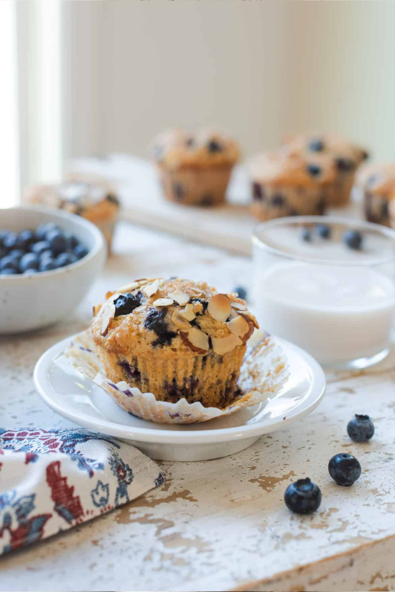 A blueberry almond muffin sits on a small white plate alongside a glass of plant-based milk