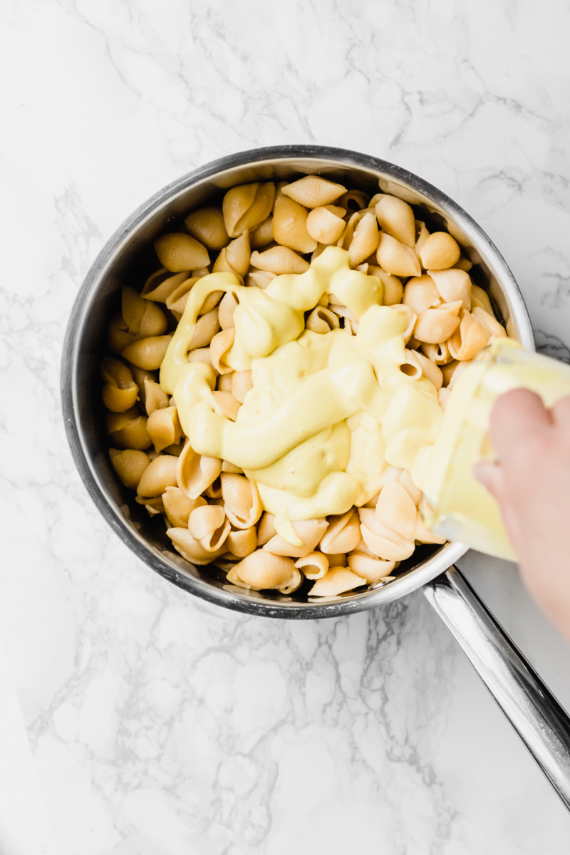 pouring yellow cheese sauce over pasta in a stainless steel pot