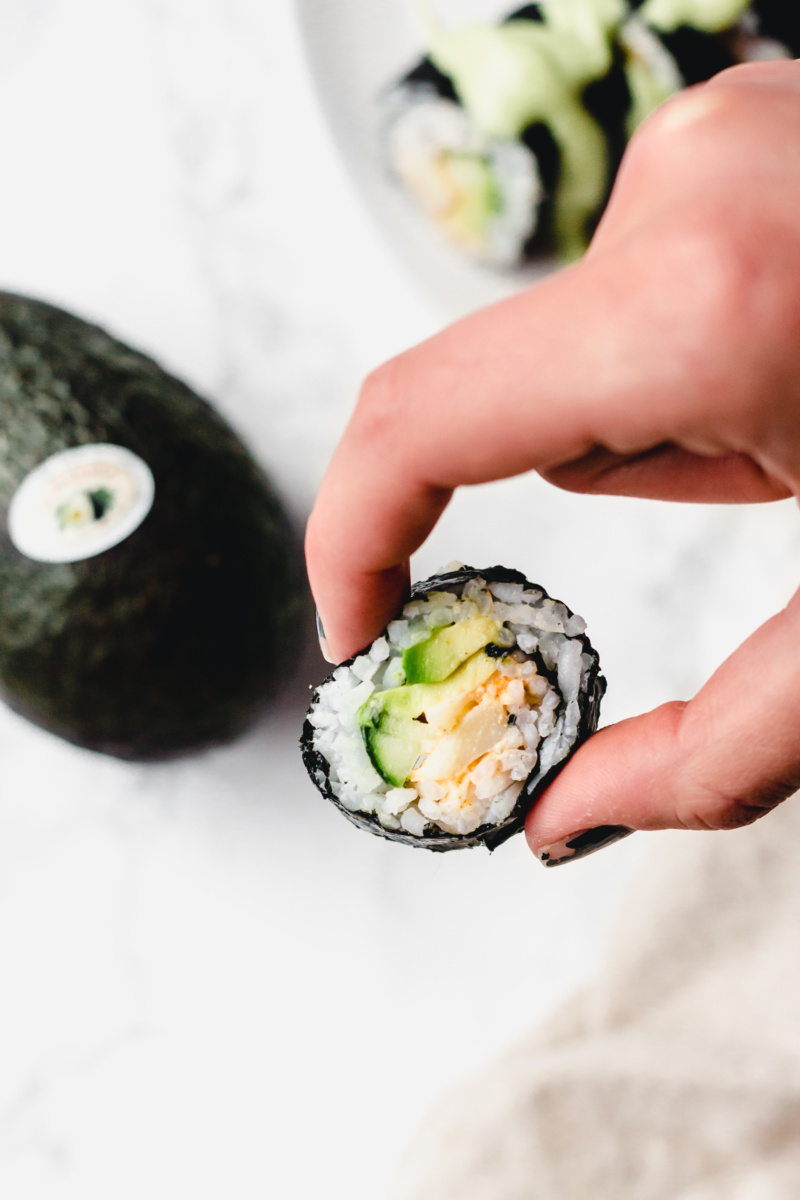 a hand holds a piece of vegan sushi, filled with avocado, cucumber and hearts of palm