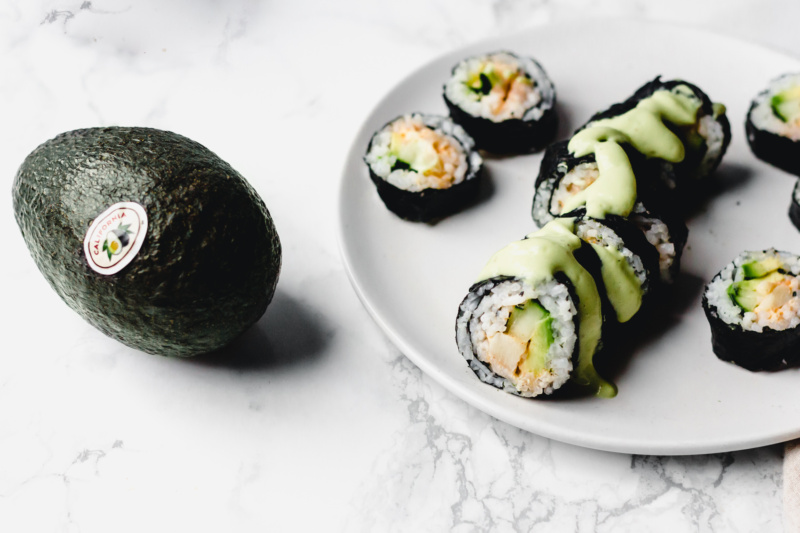 an avocado sits alongside a plate of vegan california roll slices topped with an avocado wasabi sauce