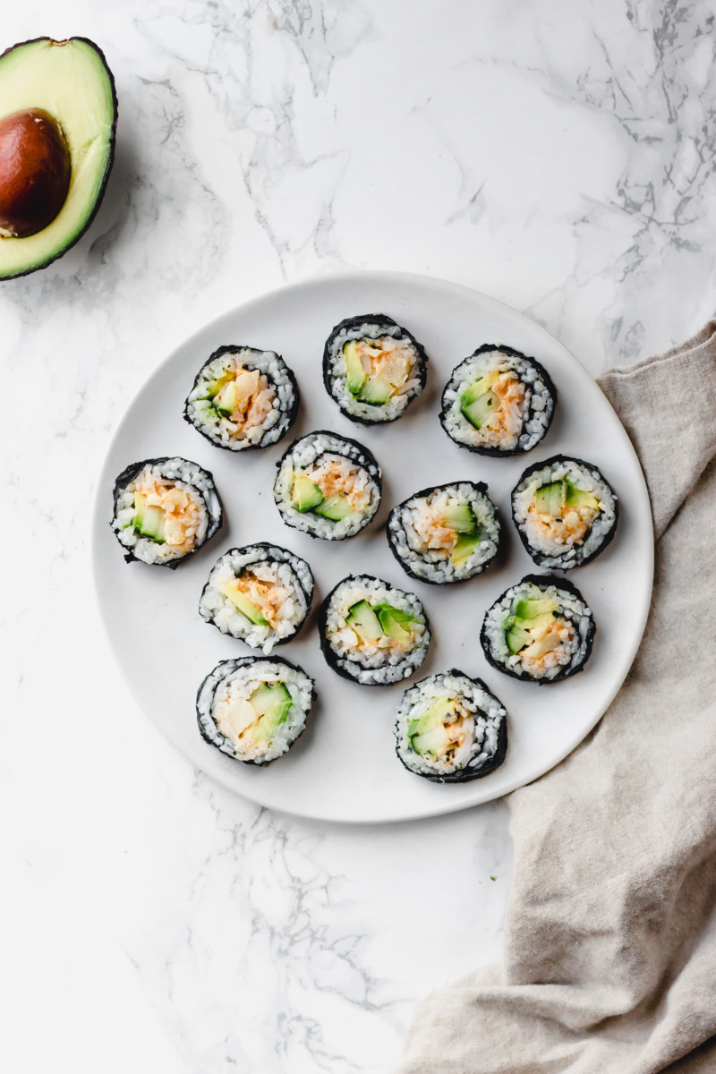 A plate of sliced vegan sushi made with cucumber, avocado and hearts of palm served alongside half of an avocado