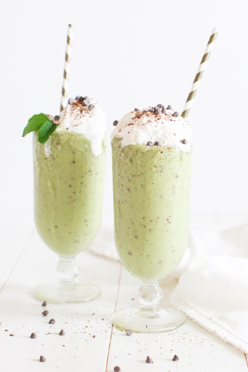 two glasses of mint chocolate chip smoothie topped with coconut whipped cream and chocolate chips