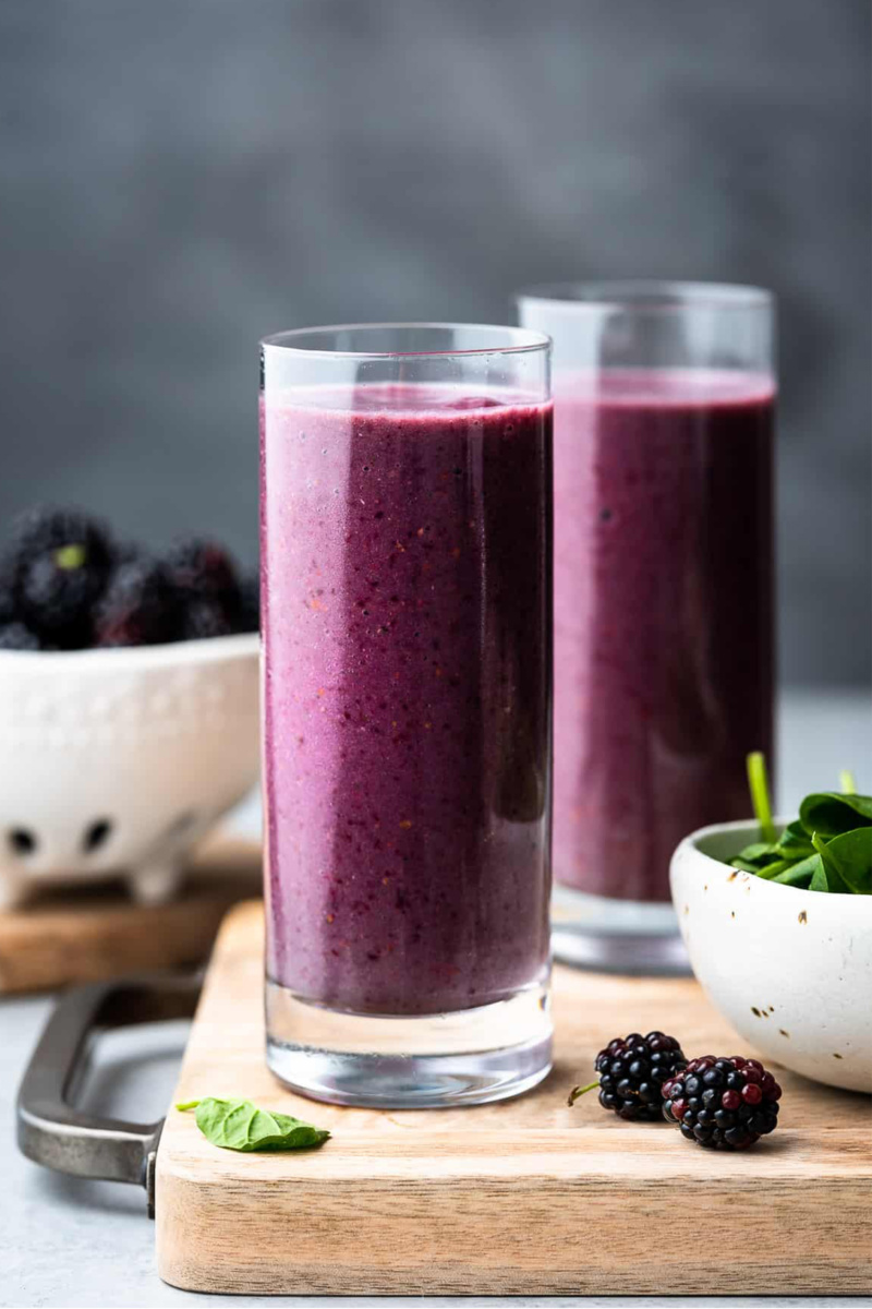 two glasses of blackberry smoothie next to a bowl of spinach