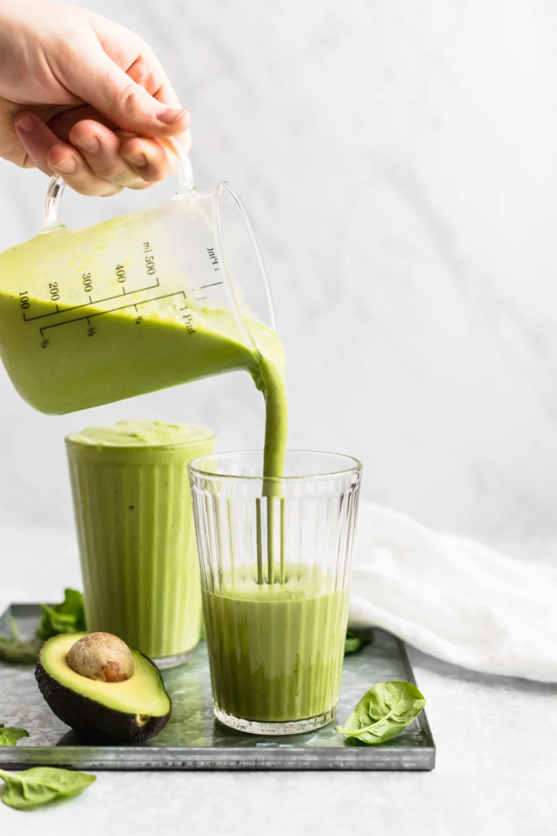 a pitcher pouring a glass of green smoothie next to a half an avocado