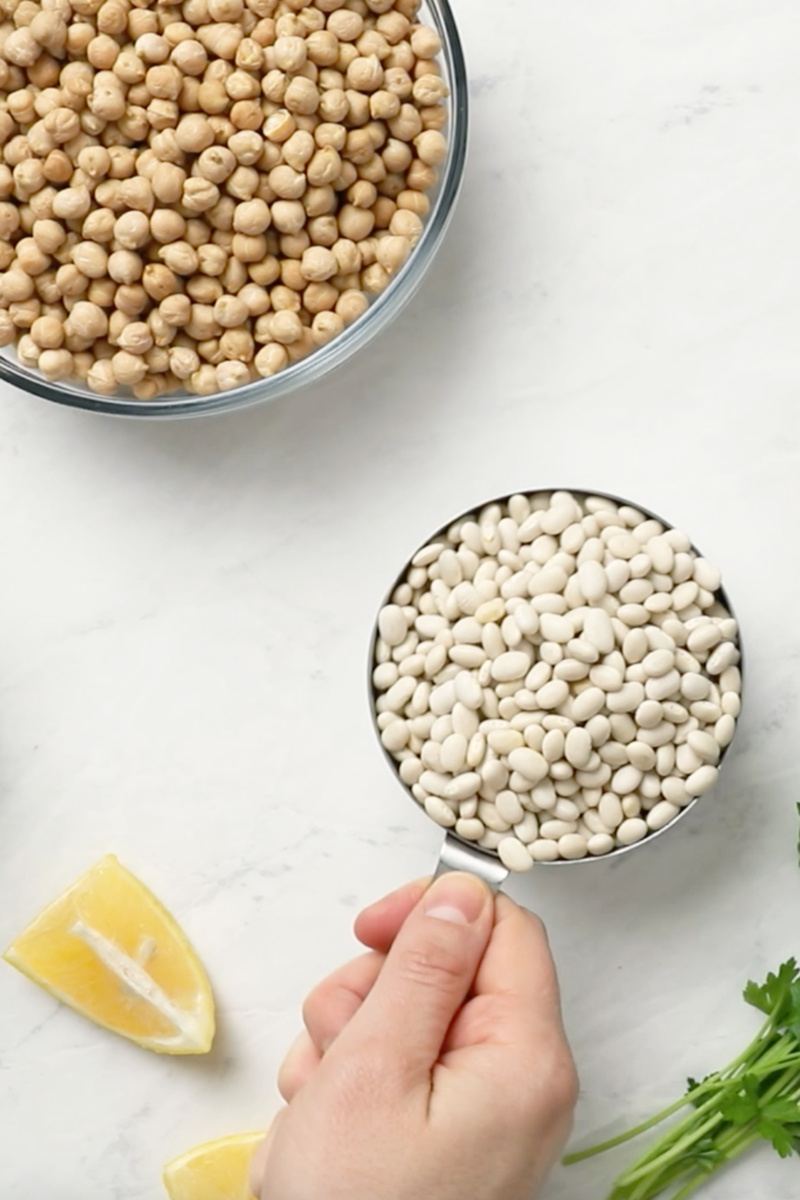 a measuring cup of dried white beans next to two bowls of lentils and chickpeas along with a wedge of lemon and a sprig of parsley