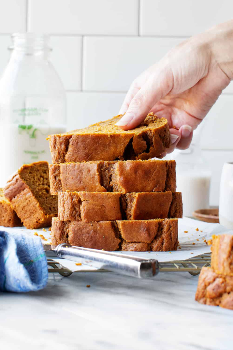 a hand grabbing a slice of pumpkin bread off of a cooling rack