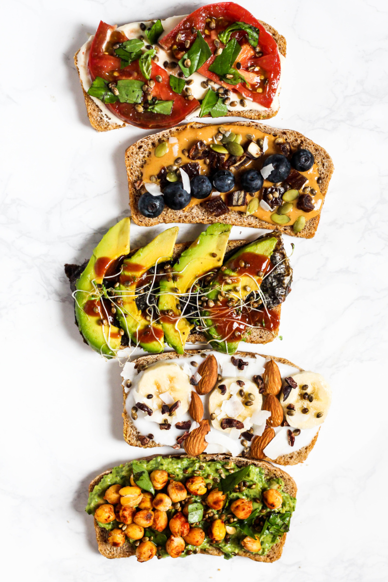 five different topped toasts, including a caprese topped toast, a peanut butter and blueberry topped toast, an avocado and hot sauce topped toast, a vegan yogurt and banana toast and a chickpea topped toast