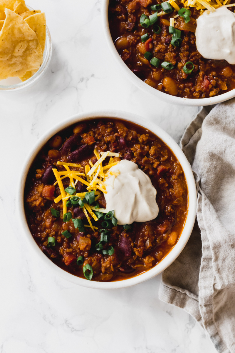 two bowls of chili served with chips and topped with sour cream, sliced green onion and cheese