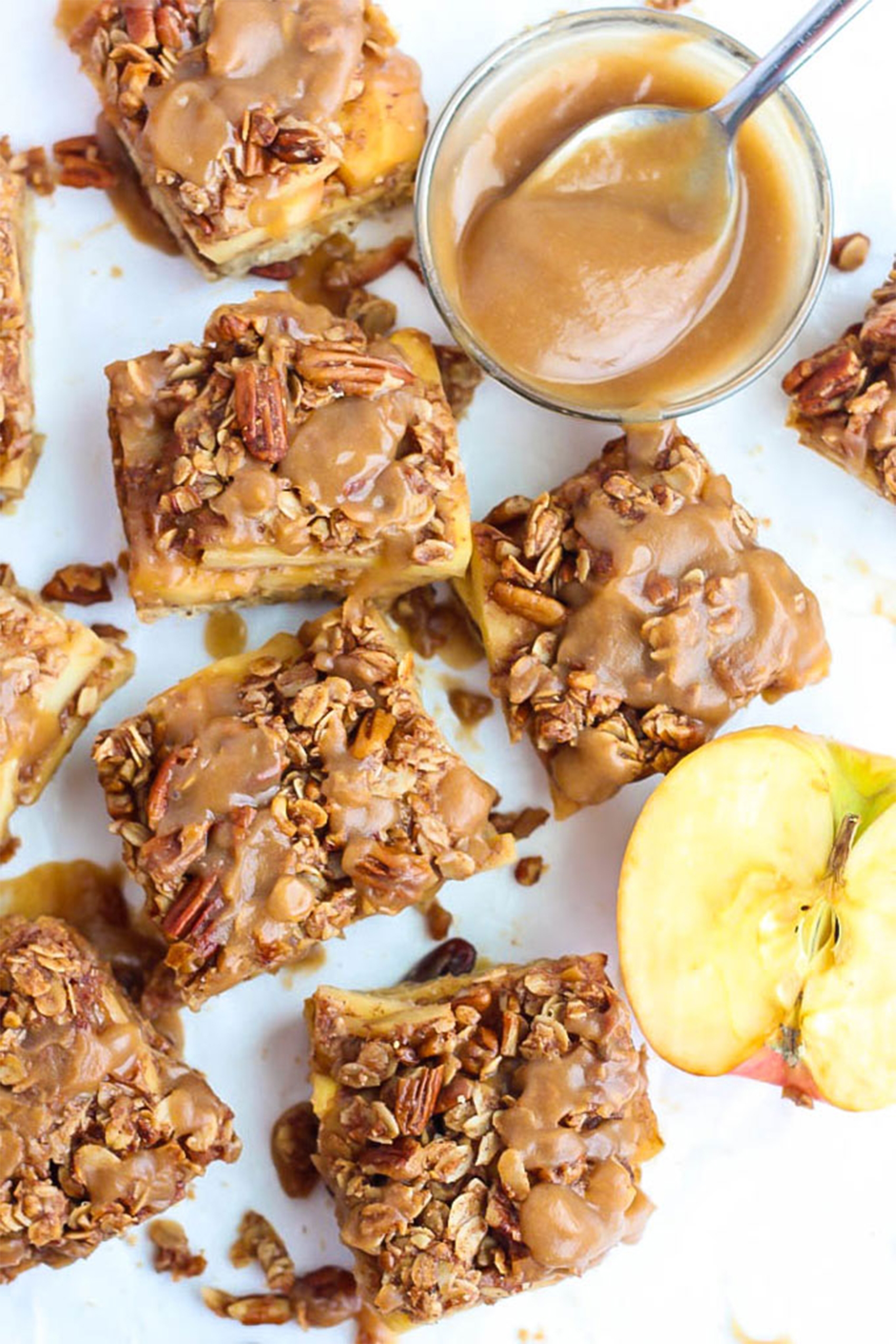 salted caramel apple pie bars that have been cut into squares sitting next to a bowl of salted caramel and a half of an apple