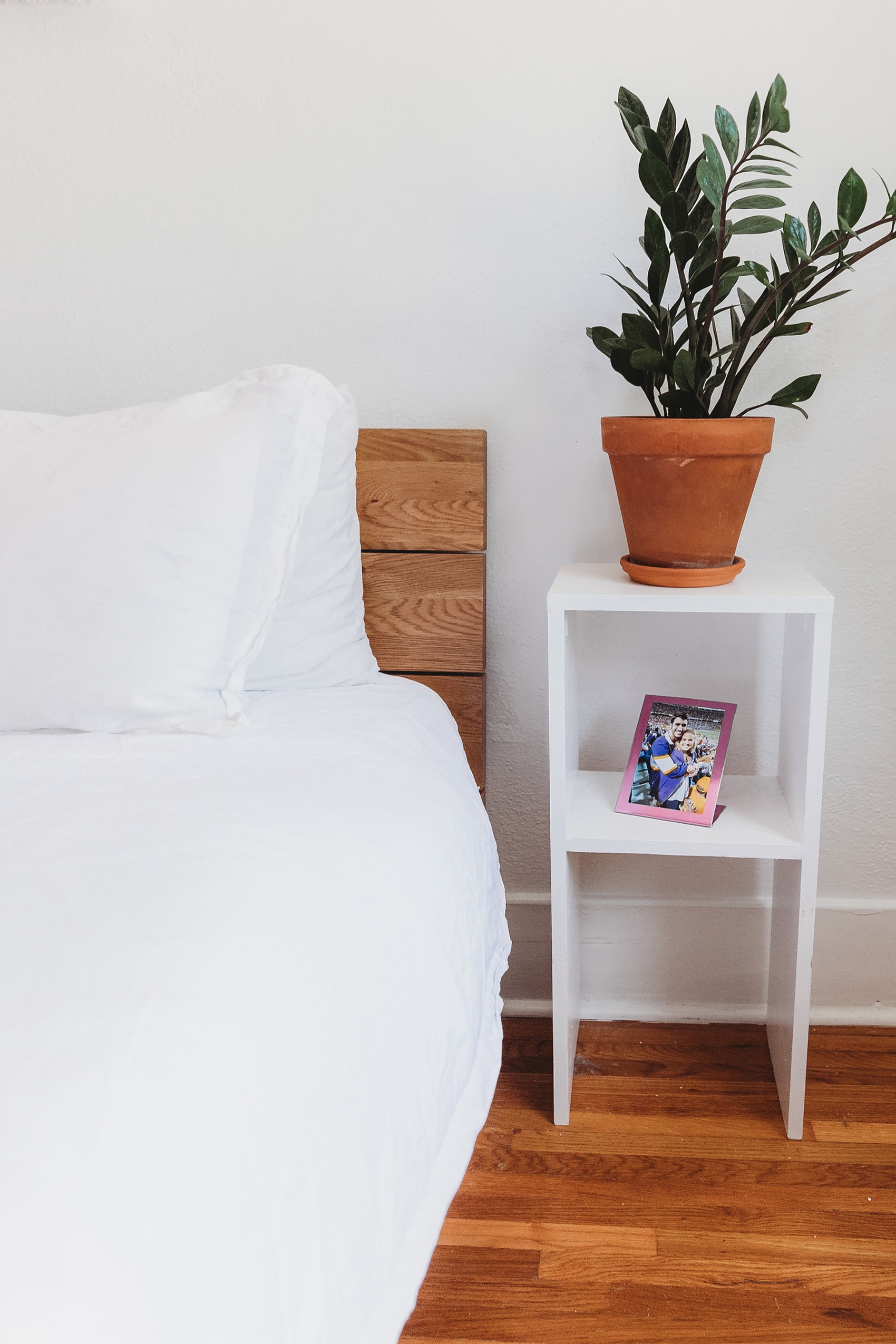 a bed made with white sheets next to a white side table topped with a plant