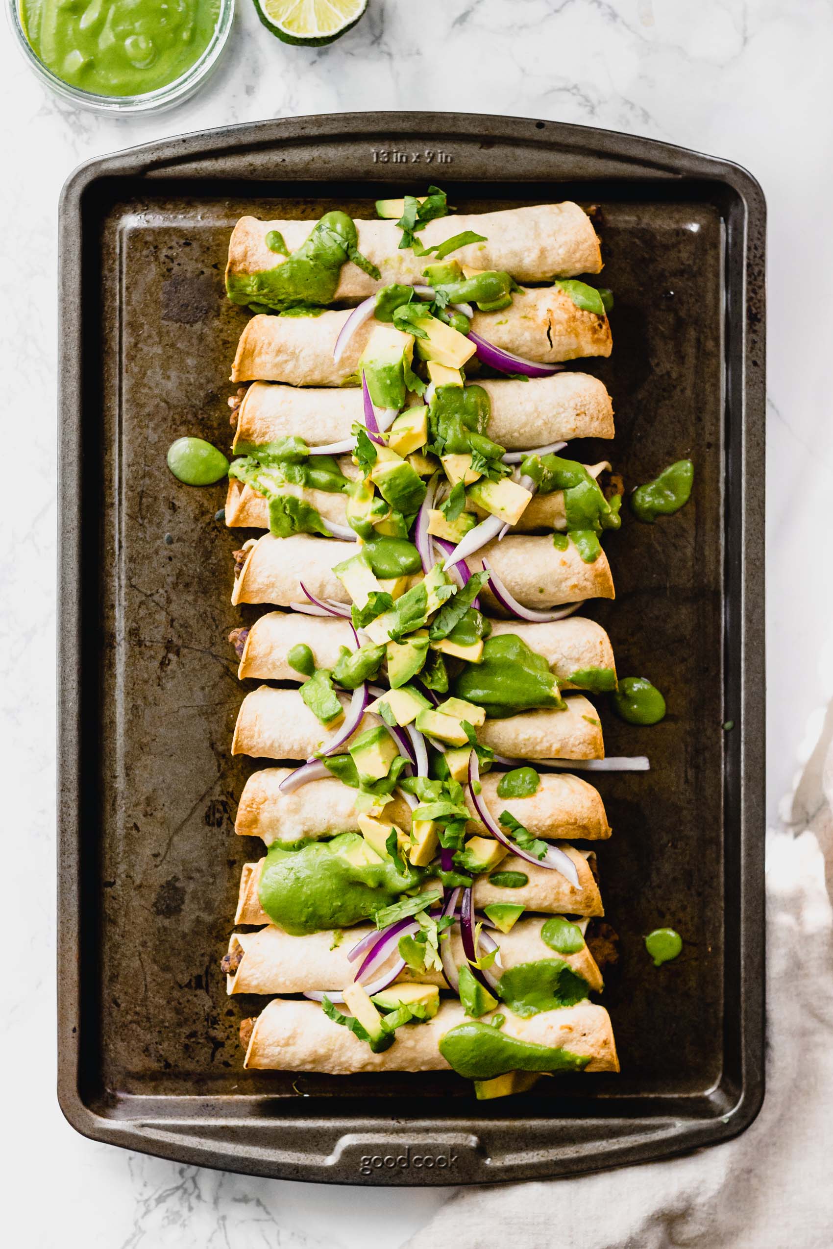 a sheet tray of vegan black bean taquitos topped with avocado, red onion and an avocado crema